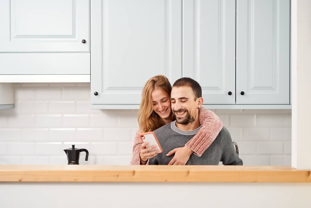Happy young couple in kitchen at home, using mobile phone - Photo, Image