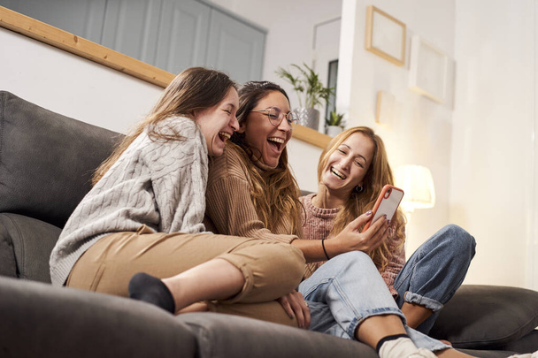Női barátok csoportja otthon a kanapén, mobiltelefonnal nevetve. - Fotó, kép