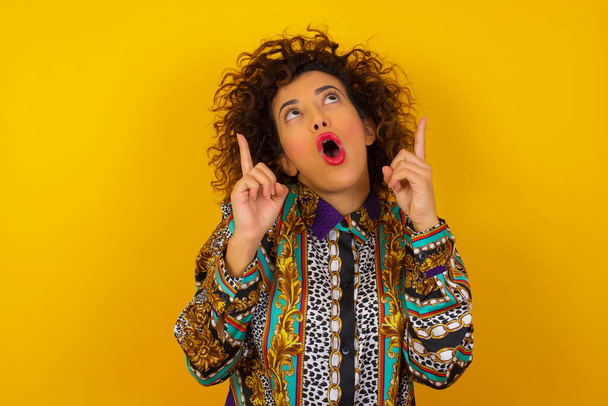 Portrait of beautiful elegant Arab woman wearing colorful shirt   over isolated background being amazed and surprised looking and pointing up with fingers showing something strange. - Photo, Image