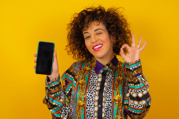 Студийный снимок возбужденной женщины, показывающей экран смартфона, моргающий глаз и делающей жест рукой. - Фото, изображение