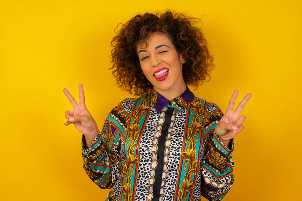Indoor portret van jonge Arabische vrouw draagt kleurrijke shirt geïsoleerd over de achtergrond met optimistische glimlach, toont vrede of overwinning gebaar met beide handen, op zoek vriendelijk. V-teken. - Foto, afbeelding