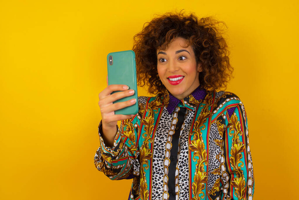 Wesoła młoda kobieta z ciemnymi włosami robi selfie z telefonem komórkowym, ubrana w luźne ubrania odizolowane na żółtym tle. - Zdjęcie, obraz