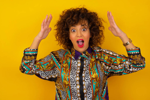 Portrait of beautiful Arab woman wearing colorful shirt  standing against yellow background, looks with excitement at camera, keeps hands raised over head, notices something unexpected. - Foto, Imagem
