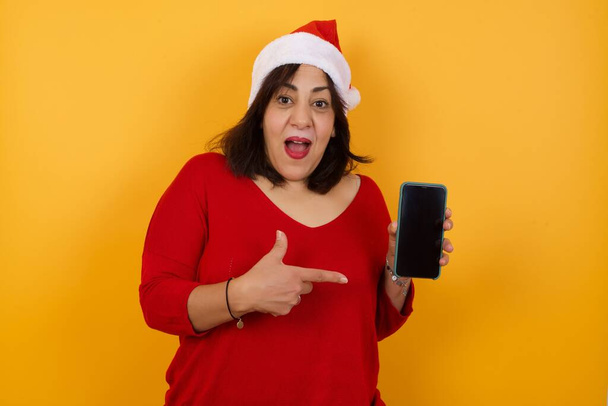 イエロースタジオのデジタルデバイスで指を指してクリスマスの帽子をかぶったエキサイティングな中年の女性 - 写真・画像