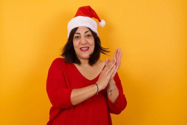 arabska kobieta w średnim wieku ubrana w świąteczny kapelusz stojąca pod żółtą ścianą, szczęśliwa, uśmiechnięta i klaszcząca w dłonie, gratulująca oklaskami. - Zdjęcie, obraz