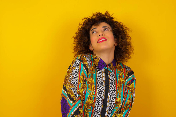 Portrait of mysterious charming Arab woman wearing colorful shirt  looking up with enigmatic smile. Beautiful smiling girl looking up standing against wall. - Fotografie, Obrázek