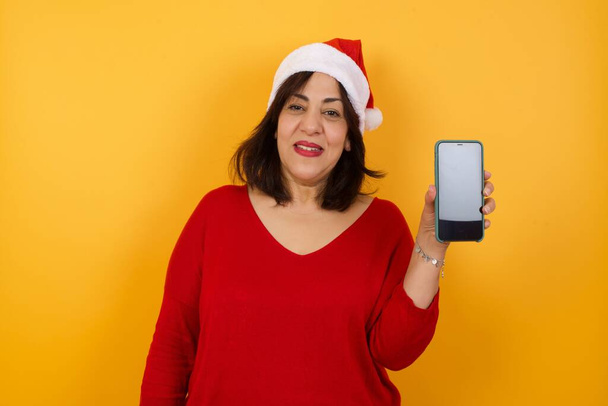 Nainen joulu hattu osoittaa älypuhelin vastaan keltainen studio tausta - Valokuva, kuva