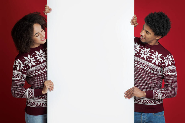 Vriendelijk Afrikaans Amerikaans jong paar in identieke kerst truien houdt blanco boord voor woorden en kijkt naar banner - Foto, afbeelding