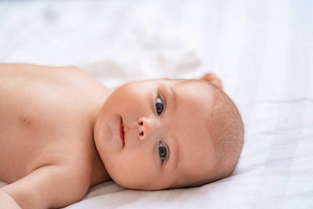 Cute baby lying on bed on white blanket and smiling. small curious child 4 months, Caucasian, lies on surface in bright cozy room, dressed in light body and looks at camera against background of room. - Фото, изображение