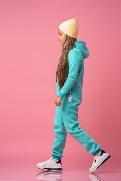 Teen girl in beanie, turquoise warm jumpsuit with hood in sneakers go on pink background studio. Full-length. Children's fashion, comfortable children's clothing for winter and autumn. Copy space - Фото, зображення
