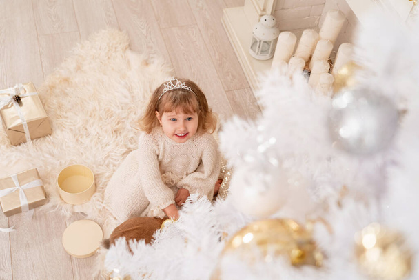 Merry Christmas and happy holidays! little girl in knitted dress and tiara sits near White Christmas tree. child looks up. Decorated living room with toys. cozy warm winter day at White house T?p view - 写真・画像