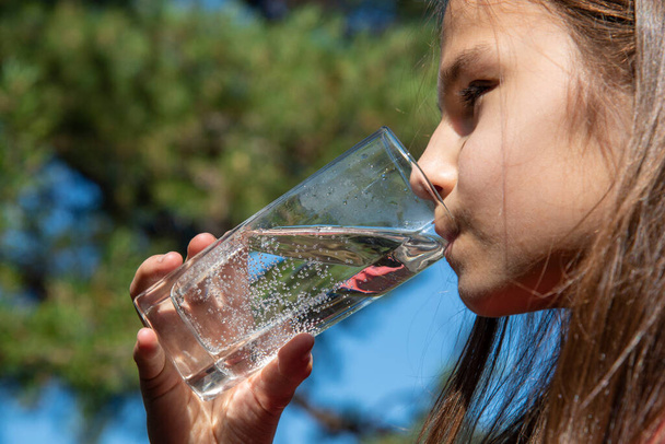 Primer plano de una niña con el pelo largo bebiendo agua clara de un vidrio transparente sobre un fondo borroso del bosque. El concepto de un estilo de vida natural saludable - Foto, imagen