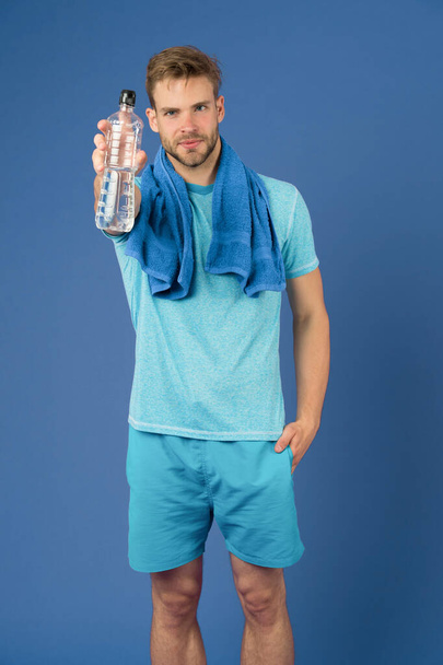Man hold water bottle on violet background. Sportsman with plastic bottle at purple wall. Athlete in blue tshirt and shorts with towel after training. Thirst and dehydration. Sport activity and energy - Photo, Image
