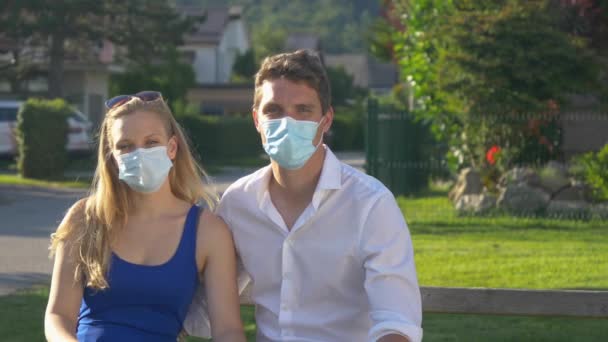 PORTRAIT Couple wearing facemasks cuddles together while sitting on a park bench - Footage, Video