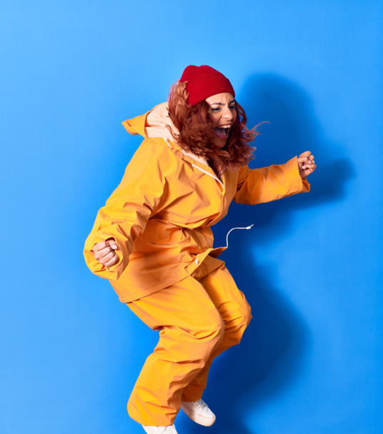 Young beautiful latin woman wearing fisher raincoat smiling happy. Jumping with smile on face celebrating with fists up over isolated blue background - Fotografie, Obrázek