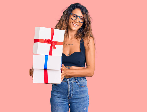 Young hispanic woman with tattoo holding gifts looking positive and happy standing and smiling with a confident smile showing teeth  - Fotografie, Obrázek