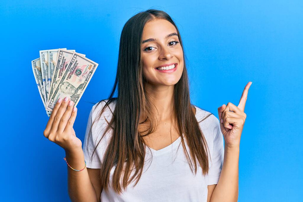 Młoda latynoska kobieta trzyma dolary uśmiechając się szczęśliwy wskazując ręką i palcem na bok  - Zdjęcie, obraz