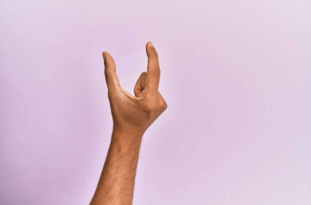 Arm und Hand eines kaukasischen jungen Mannes über rosa isoliertem Hintergrund, der unsichtbare Dinge pflückt und nimmt, mit Fingern Gegenstände hält, die Raum zeigen  - Foto, Bild