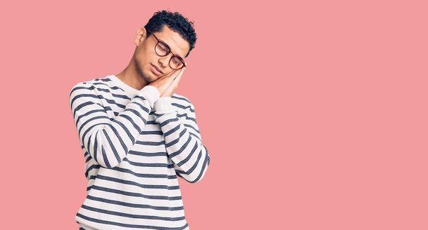 Hispanic handsome young man wearing casual clothes and glasses sleeping tired dreaming and posing with hands together while smiling with closed eyes.  - 写真・画像