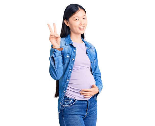 Młoda piękna Chinka w ciąży spodziewa się dziecka uśmiechniętego szczęśliwą twarzą mrugającego do kamery robiącego znak zwycięstwa palcami. numer dwa..  - Zdjęcie, obraz