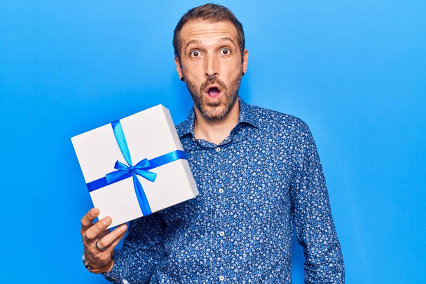 Young handsome man holding gift scared and amazed with open mouth for surprise, disbelief face  - Fotoğraf, Görsel
