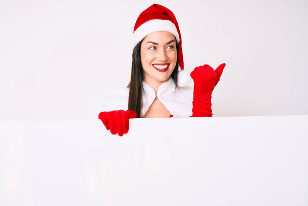 Jovem mulher vestindo traje de Papai Noel segurando banner vazio apontando polegar até o lado sorrindo feliz com a boca aberta  - Foto, Imagem