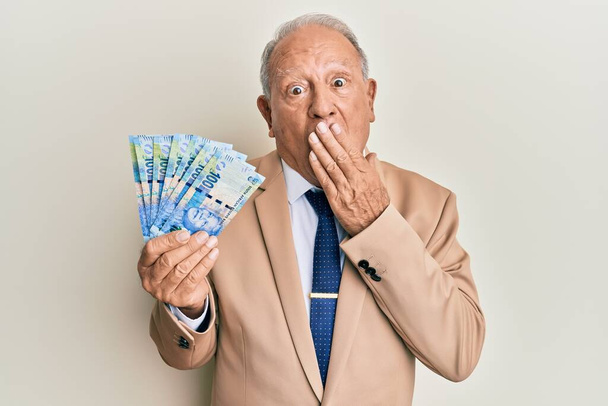 Hombre caucásico mayor sosteniendo billetes sudafricanos de 20 rand cubriendo la boca con la mano, sorprendido y temeroso de equivocarse. expresión sorprendida  - Foto, Imagen