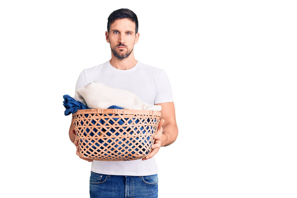 Young handsome man holding laundry basket thinking attitude and sober expression looking self confident  - Foto, Imagem