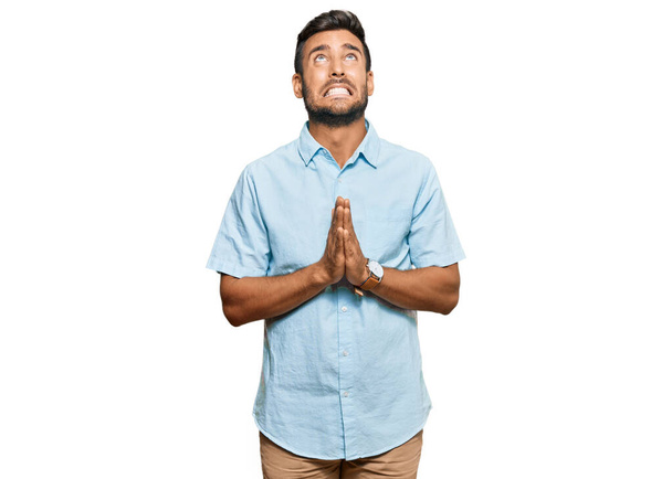 Красивий іспаномовний чоловік в повсякденному одязі молиться і молиться руками разом з надією висловлюватися на обличчі дуже емоційно і стурбовано. благання
.  - Фото, зображення