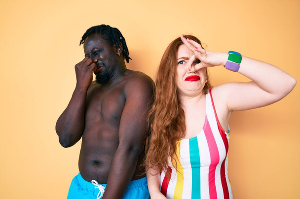 Interracial couple wearing swimwear smelling something stinky and disgusting, intolerable smell, holding breath with fingers on nose. bad smell  - Fotoğraf, Görsel