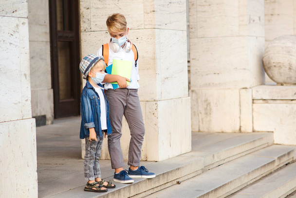 School kids wearing face mask during coronavirus outbreak. Children going home after school. Coronavirus quarantine and lockdown. Cute brothers in medical masks walking at street. - Photo, Image