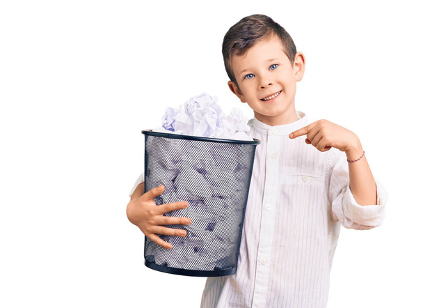 Lindo niño rubio sosteniendo papelera llena de papeles arrugados sonriendo feliz señalando con la mano y el dedo  - Foto, imagen