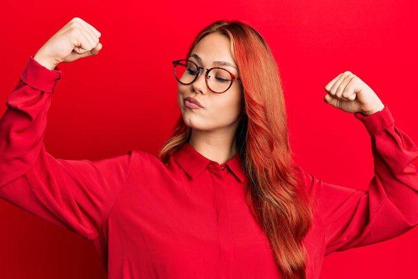 Joven mujer pelirroja hermosa con ropa casual y gafas sobre fondo rojo mostrando los músculos de los brazos sonriendo orgulloso. concepto de fitness.  - Foto, Imagen