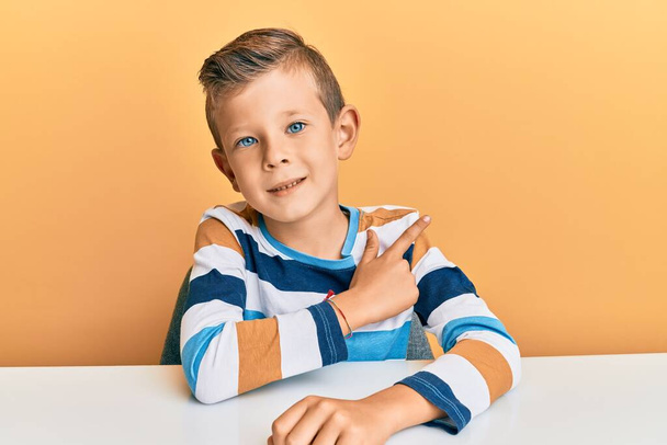 Adorabile bambino caucasico che indossa abiti casual seduto sul tavolo allegro con un sorriso sul viso che punta con mano e dito verso il lato con espressione felice e naturale  - Foto, immagini