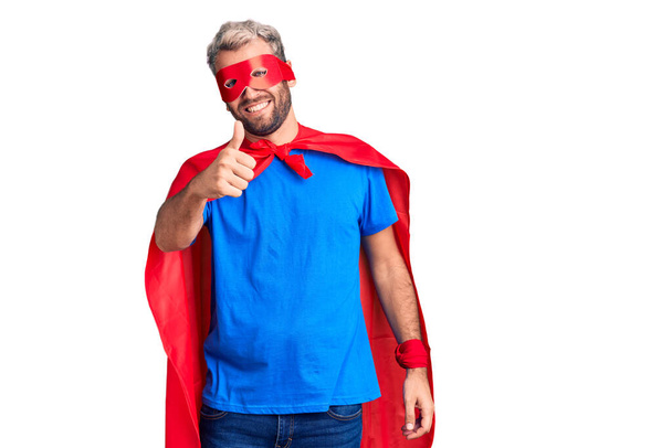 Young blond man wearing super hero custome doing happy thumbs up gesture with hand. approving expression looking at the camera showing success.  - Zdjęcie, obraz