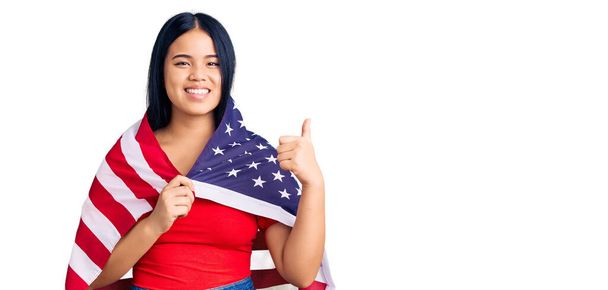 Jeune belle fille asiatique tenant drapeau des États-Unis souriant heureux et positif, pouce levé faire excellent et signe d'approbation  - Photo, image