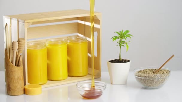 Três jarros de mel CBD colocados em uma caixa de madeira. Ao redor estão uma planta de maconha em um vaso branco, uma tigela com sementes de cânhamo, com ênfase no mel pingando em uma tigela de vidro pequena. - Filmagem, Vídeo