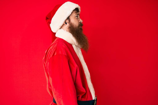Όμορφος νεαρός κόκκινο κεφάλι άνθρωπος με μακριά γενειάδα φορώντας κοστούμι Santa Claus ψάχνει για πλευρά, χαλαρώστε προφίλ ποζάρουν με φυσικό πρόσωπο με αυτοπεποίθηση χαμόγελο.  - Φωτογραφία, εικόνα
