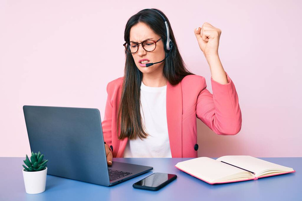 Young caucasian woman sitting at the desk wearing call center agent headset working using laptop annoyed and frustrated shouting with anger, yelling crazy with anger and hand raised  - 写真・画像