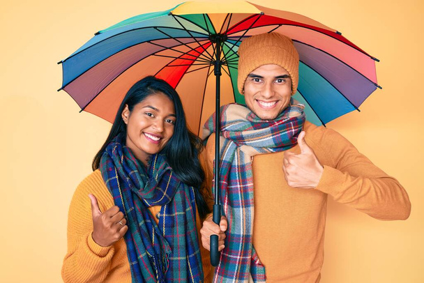 Piękna latynoska młoda para pod kolorowym parasolem uśmiechnięta szczęśliwa i pozytywna, kciuk do góry robi doskonałe i znak aprobaty  - Zdjęcie, obraz