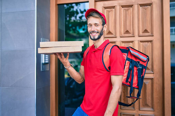 Blanc livraison homme portant uniforme rouge et livraison sac à dos souriant heureux à l'extérieur tenant boîte à pizza - Photo, image