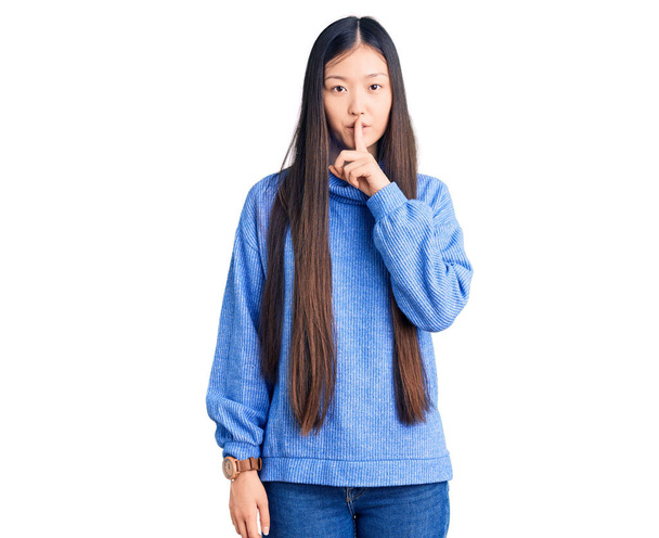 唇に指で静かにするように求めるカジュアルなタートルネックセーターを着て若い美しい中国の女性。沈黙と秘密の概念.  - 写真・画像