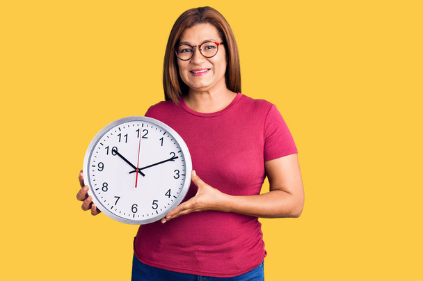 Latynoska kobieta w średnim wieku trzyma duży zegar patrząc pozytywnie i szczęśliwy stojąc i uśmiechając się z ufnym uśmiechem pokazując zęby  - Zdjęcie, obraz