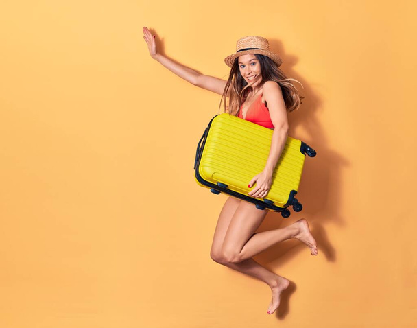 Young beautiful woman on vacation wearing bikini and hat smiling happy. Jumping with smile on face holding cabin bag over isolated background - Photo, Image