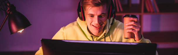 Jugador alegre en auriculares sosteniendo café para ir y mirando el monitor de la computadora en primer plano borroso, bandera  - Foto, Imagen