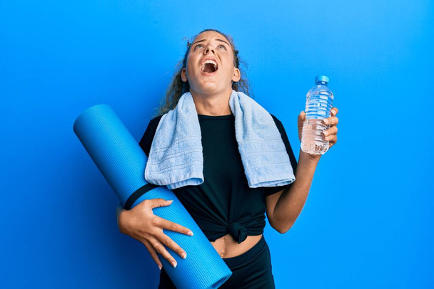 Beautiful caucasian teenager girl holding yoga mat and water bottle angry and mad screaming frustrated and furious, shouting with anger looking up.  - 写真・画像