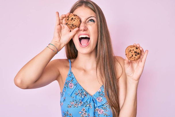 Young blonde girl holding cookie angry and mad screaming frustrated and furious, shouting with anger looking up.  - 写真・画像