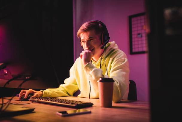 Positive gamer in headset using computer near smartphone and takeaway coffee on blurred foreground  - Foto, afbeelding