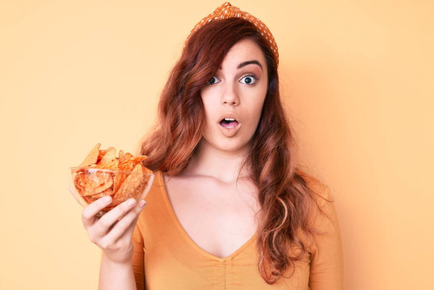 Joven hermosa mujer sosteniendo nachos patatas fritas asustado y sorprendido con la boca abierta para sorpresa, cara de incredulidad  - Foto, imagen