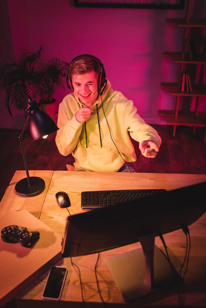 KYIV, UKRAINE - AUGUST 21, 2020: Smiling gamer in headset pointing with fingers at computer near joystick, pizza box and smartphone on blurred foreground  - Фото, зображення